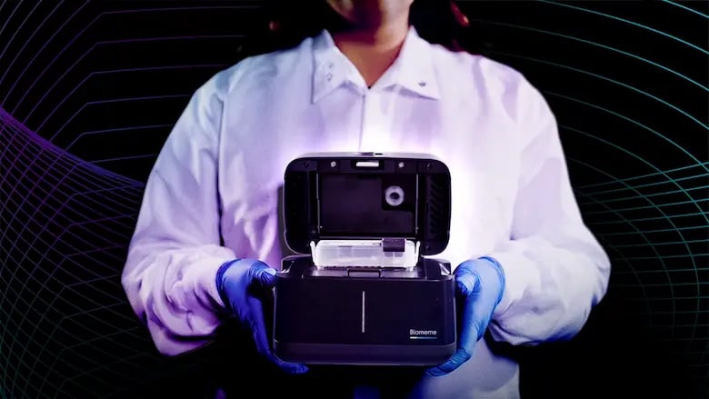 Scientist holding a Biomeme test and platform on dark background representing One Health wearing blue gloves and a white shirt. 